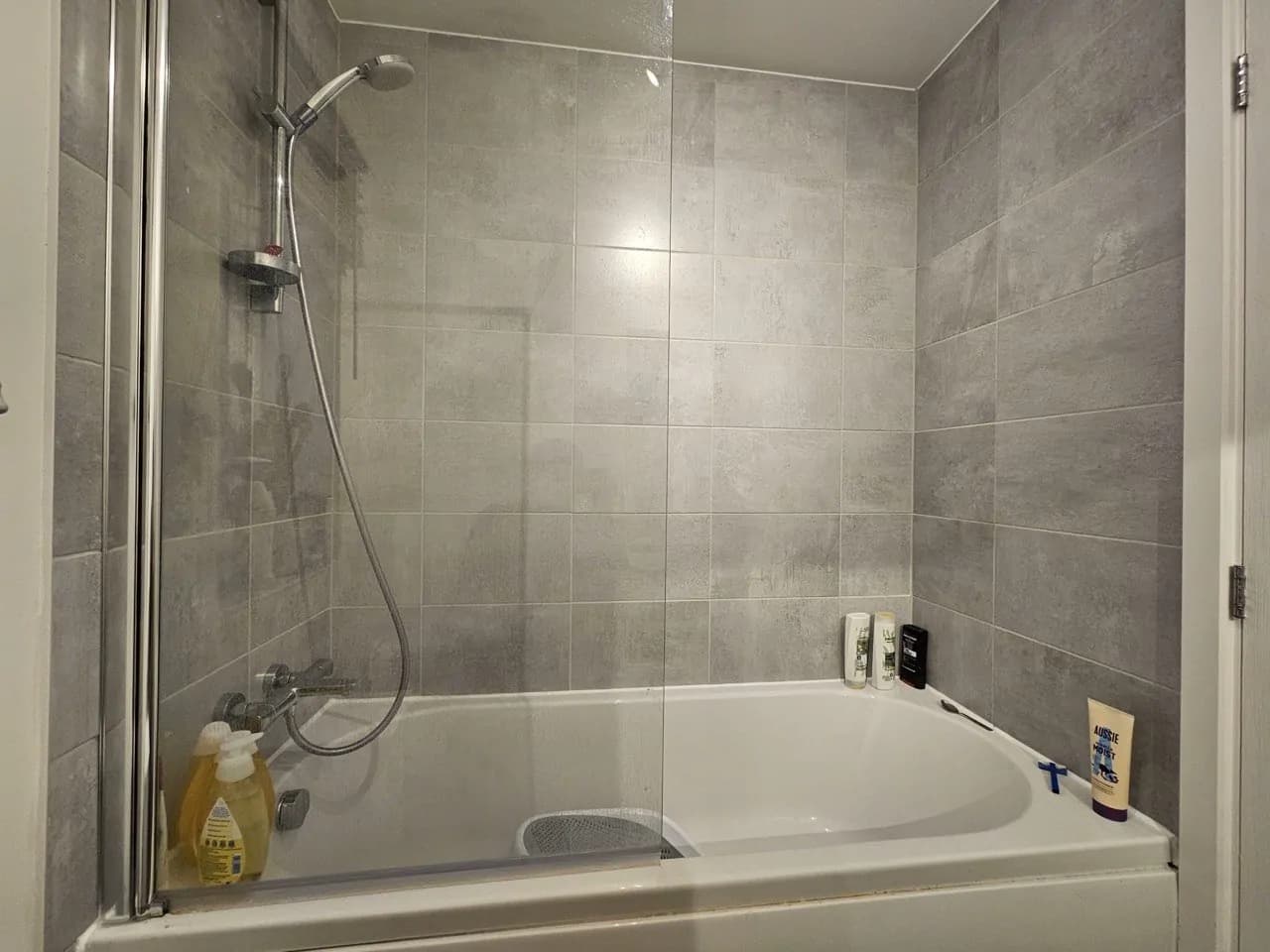 A finished bath with a glass screen, bar shower and grey wall tiles