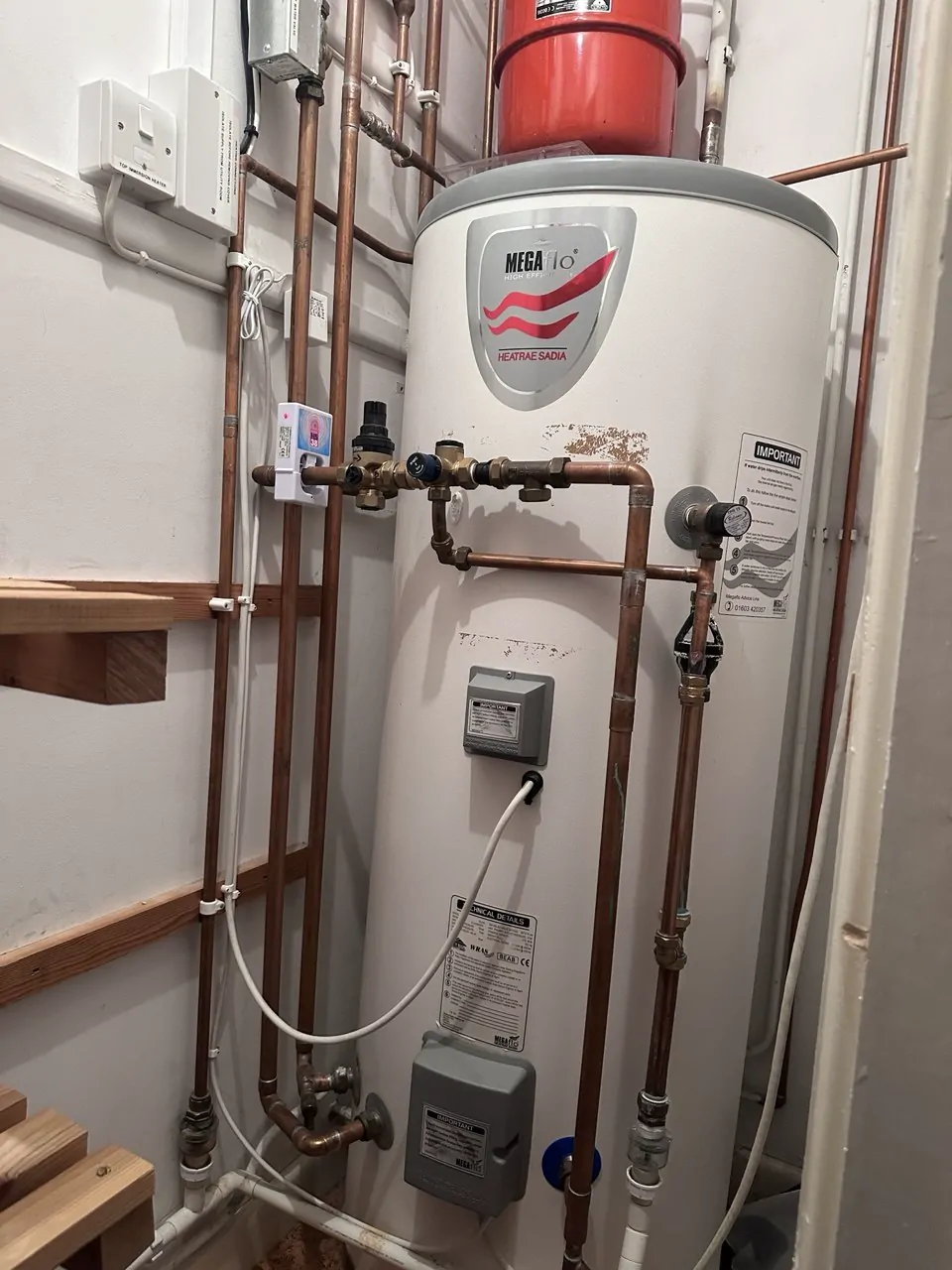A white hot water cylinder in an airing cupboard with copper pipework and valves