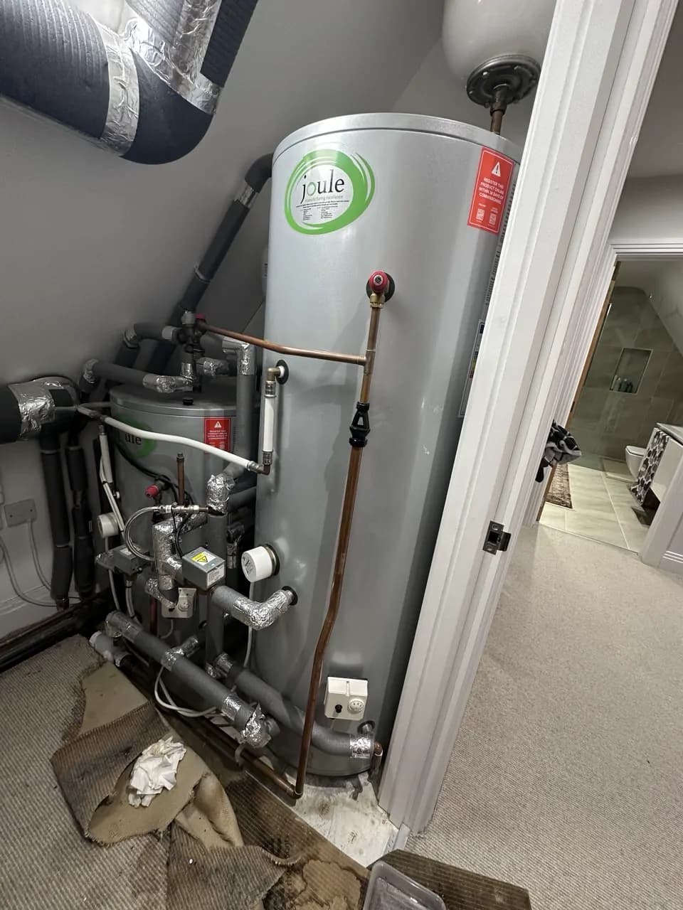 Two grey hot water cylinders with insulated pipes in a loft cupboard