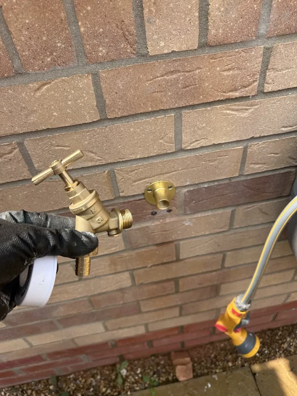 A gloved hand holding a new brass outside tap next to its wall plate on a brick wall