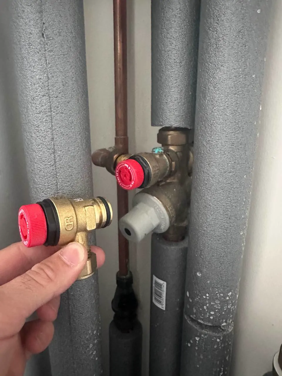 A hand holding a new brass safety valve next to the one it replaces on insulated pipework