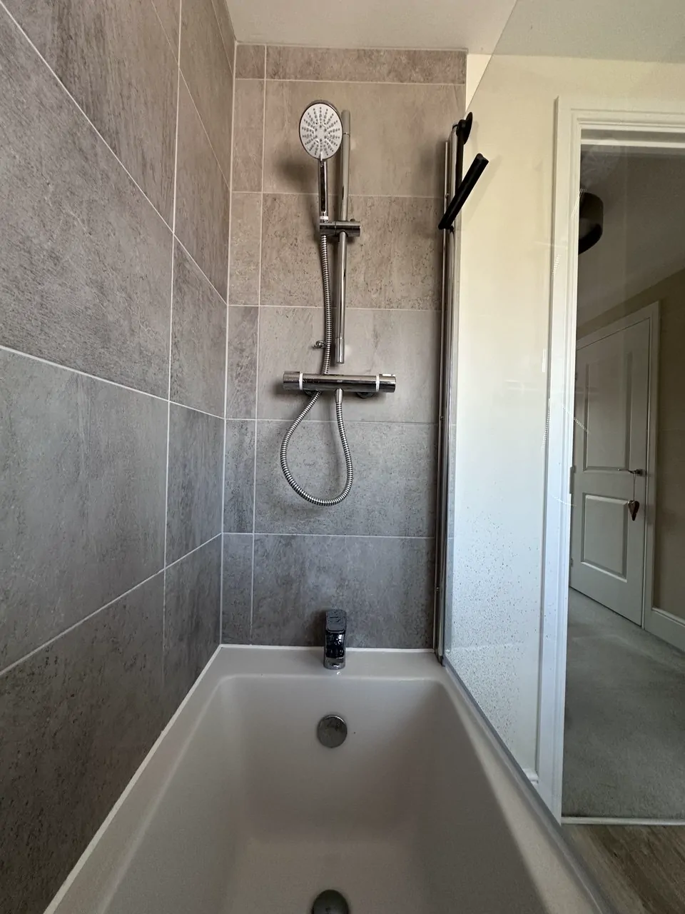 A chrome bar shower and riser rail fitted above a new bath with grey tiled walls
