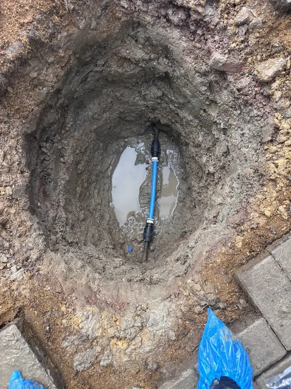 A new length of blue water supply pipe joined into the old pipe at the bottom of a hand-dug hole